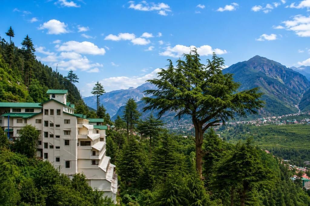 A multi-story building with a green roof, surrounded by dense forest and mountains in the background.