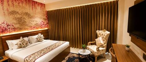 Quarto de hotel moderno com uma cama grande, um espaço de estar com uma cadeira e mesa, uma televisão e um detalhe decorativo na parede.
