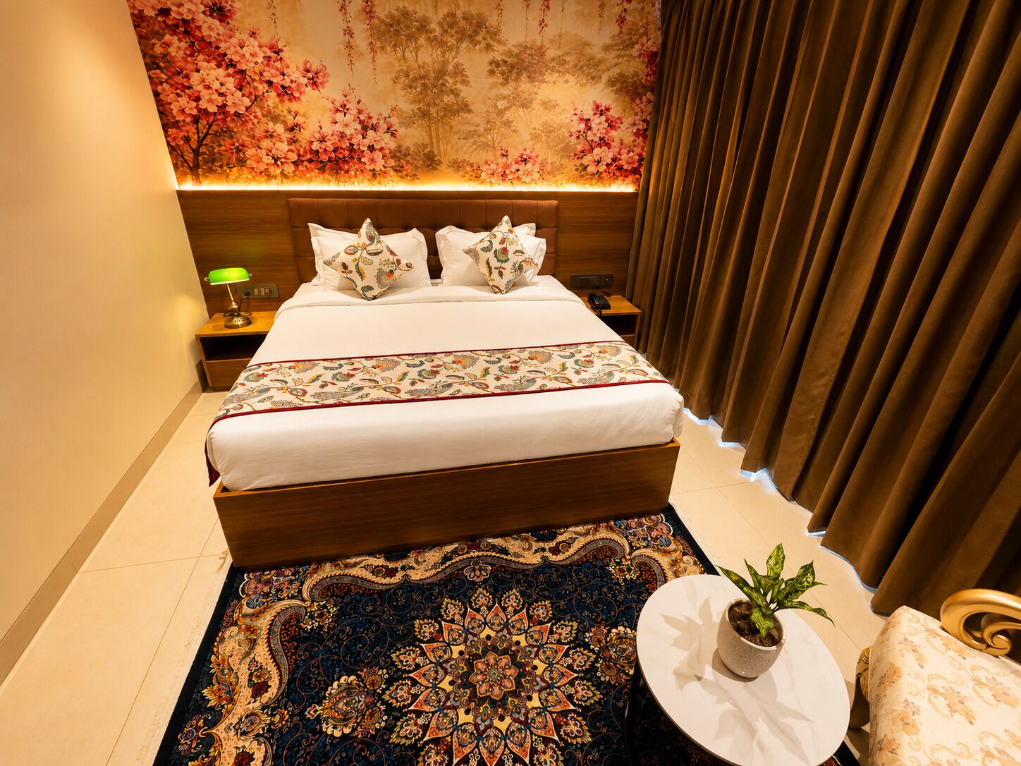 A hotel room with a large bed, a nightstand with a lamp, a round table with a plant, and a patterned rug.