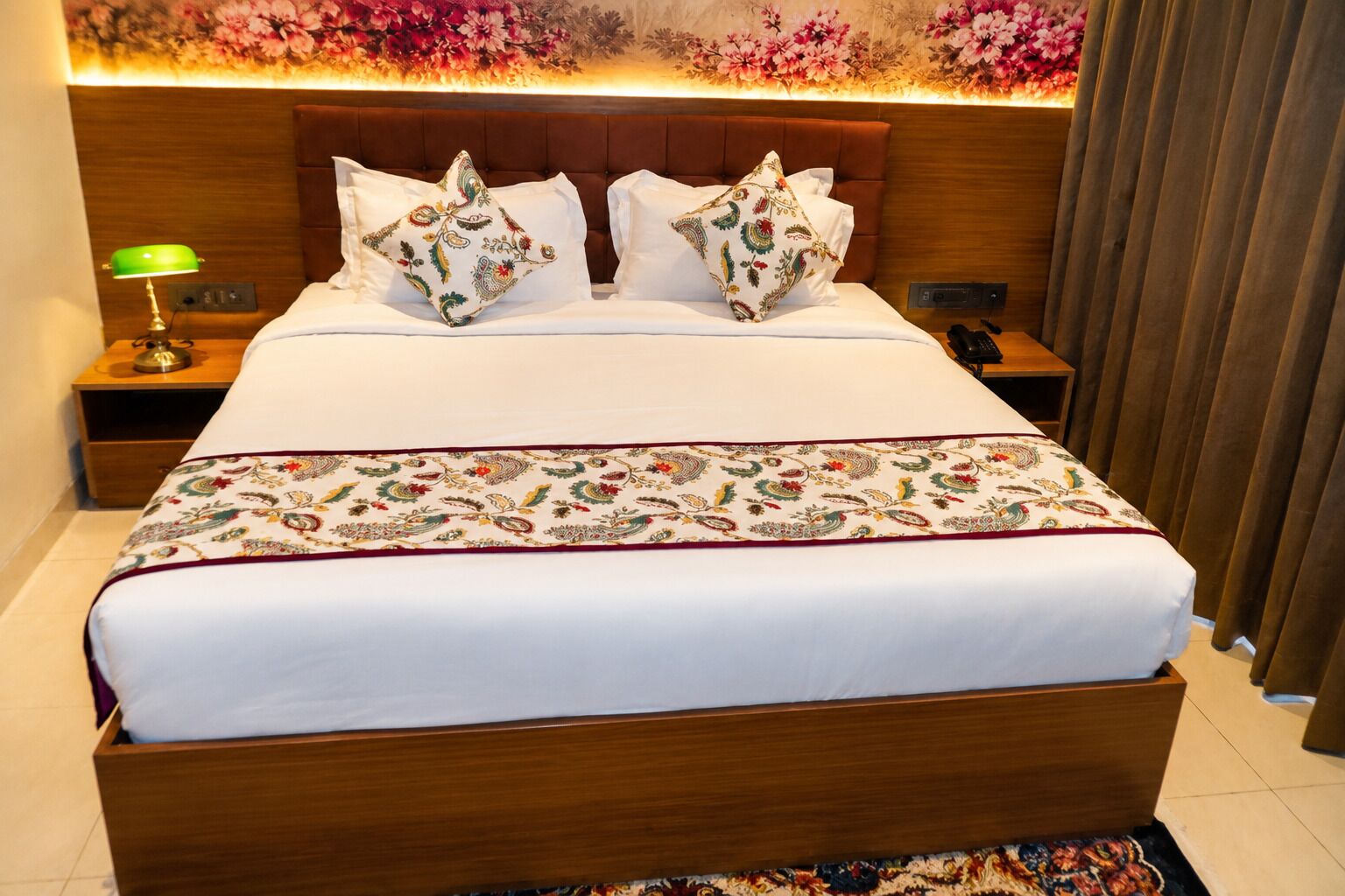 Une chambre d’hôtel avec un grand lit, deux tables de chevet, une lampe de bureau verte et une tête de lit à motifs floraux.