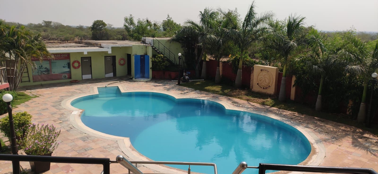 Une piscine à bord incurvé, entourée d’une zone pavée avec des marches menant à un bâtiment à la façade verdoyante et sur lequel est apposée une enseigne.