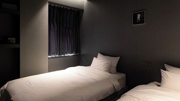 Quarto com duas camas, uma janela com cortinas, um quadro pequeno na parede e uma prateleira com objetos.