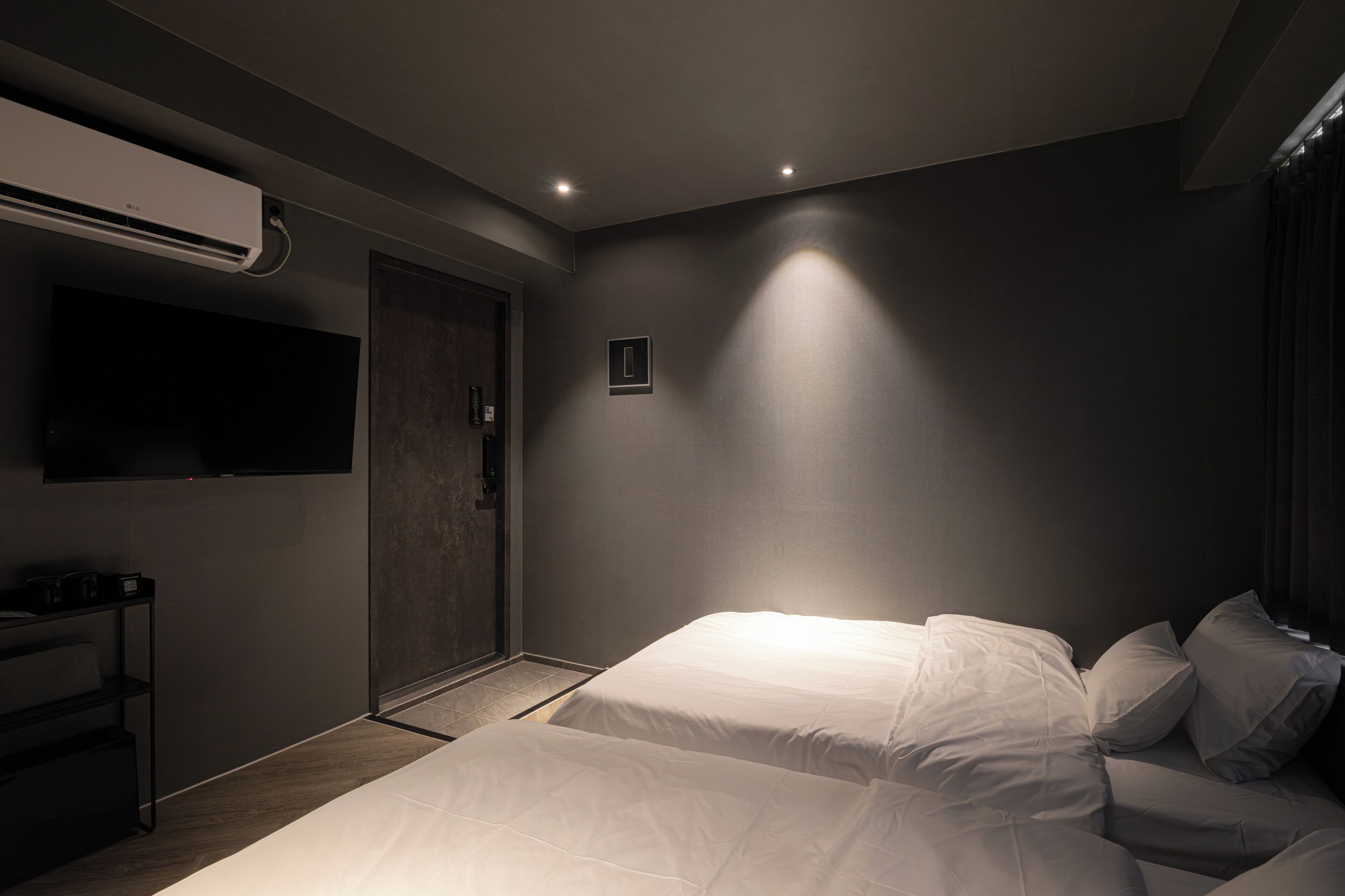 A modern hotel room with a bed, bedside table, TV, air conditioning unit, and a door.