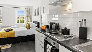 A compact hotel room with a kitchenette, a bed with a yellow blanket, a window with a view, and a door with a curtain. - Cheshire Gold: Beautiful studio apartment near  Warrington town centre (Warrington)