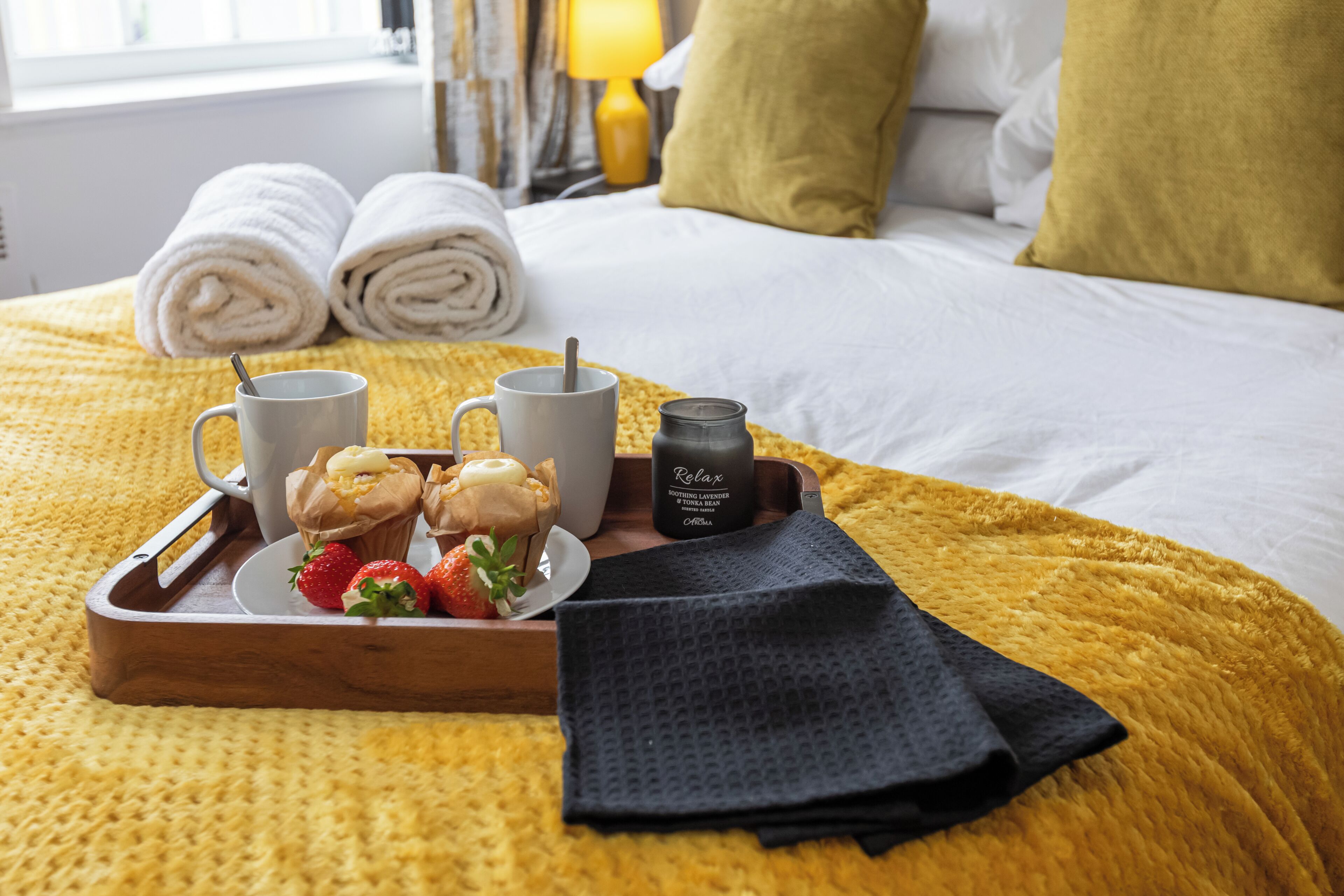 En pænt redt seng med sammenrullede håndklæder, en bakke med muffins, jordbær og et Relax-lys.
