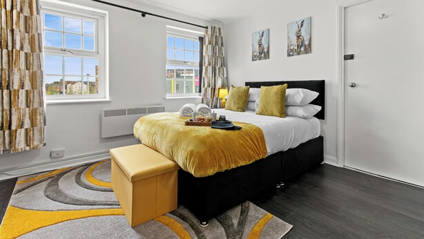 A modern bedroom with a bed, bedside tables, a bench, a window, and artwork on the wall. - Cheshire Gold: Beautiful studio apartment near  Warrington town centre (Warrington)