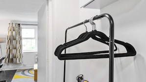 A metal rack with hangers, a vacuum cleaner, and a patterned rug in a room with a window and a curtain. - Cheshire Gold: Beautiful studio apartment near  Warrington town centre (Warrington)