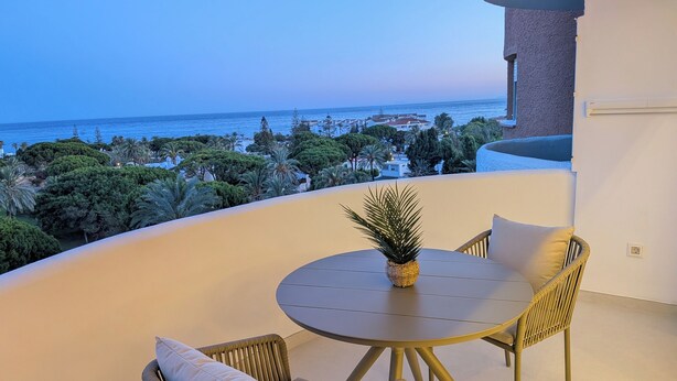 Ein Balkon mit rundem Tisch und Korbstühlen, der auf eine Küstenlandschaft mit Bäumen und Gebäuden blickt.