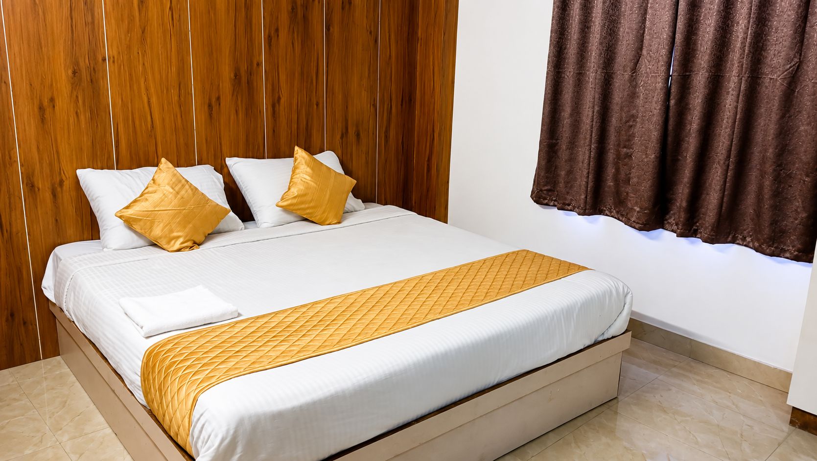 Una cama bien tendida con ropa de cama blanca y un detalle dorado, un cabecero de madera y una cortina marrón.