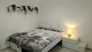 Ett sovrum med en säng, ett nattduksbord med en lampa, en växt som hänger på väggen och ett vitt litet bord.