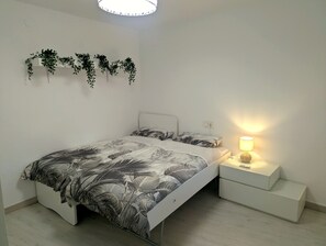 Ein Schlafzimmer mit einem Bett, einem Nachttisch mit Lampe, einer Wandpflanze und einem weißen Beistelltisch.