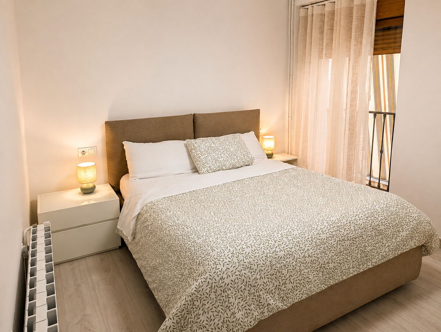 A bedroom with a bed, two bedside lamps, a radiator, and a window with curtains.