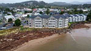 A multi-story hotel with a flat roof, located near a beach with driftwood and a stone wall.
