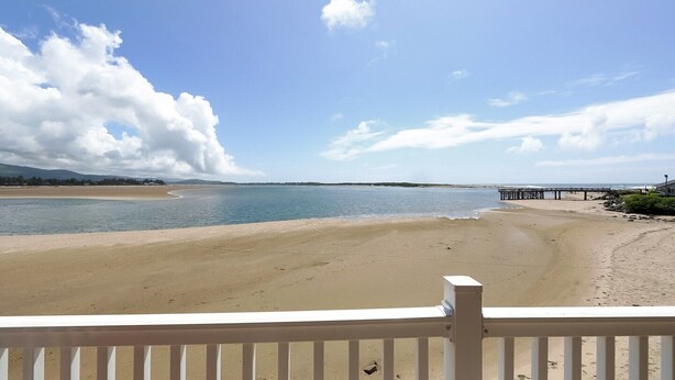 A beach with a pier extending into the water, a clear sky, and a wooden deck in the foreground.