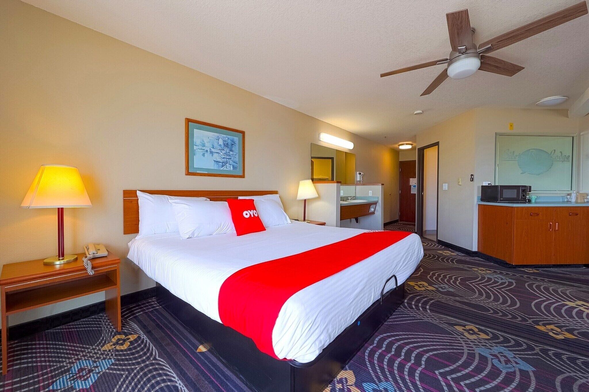 A hotel room with a bed, a desk, a mirror, a ceiling fan, and a bathroom.