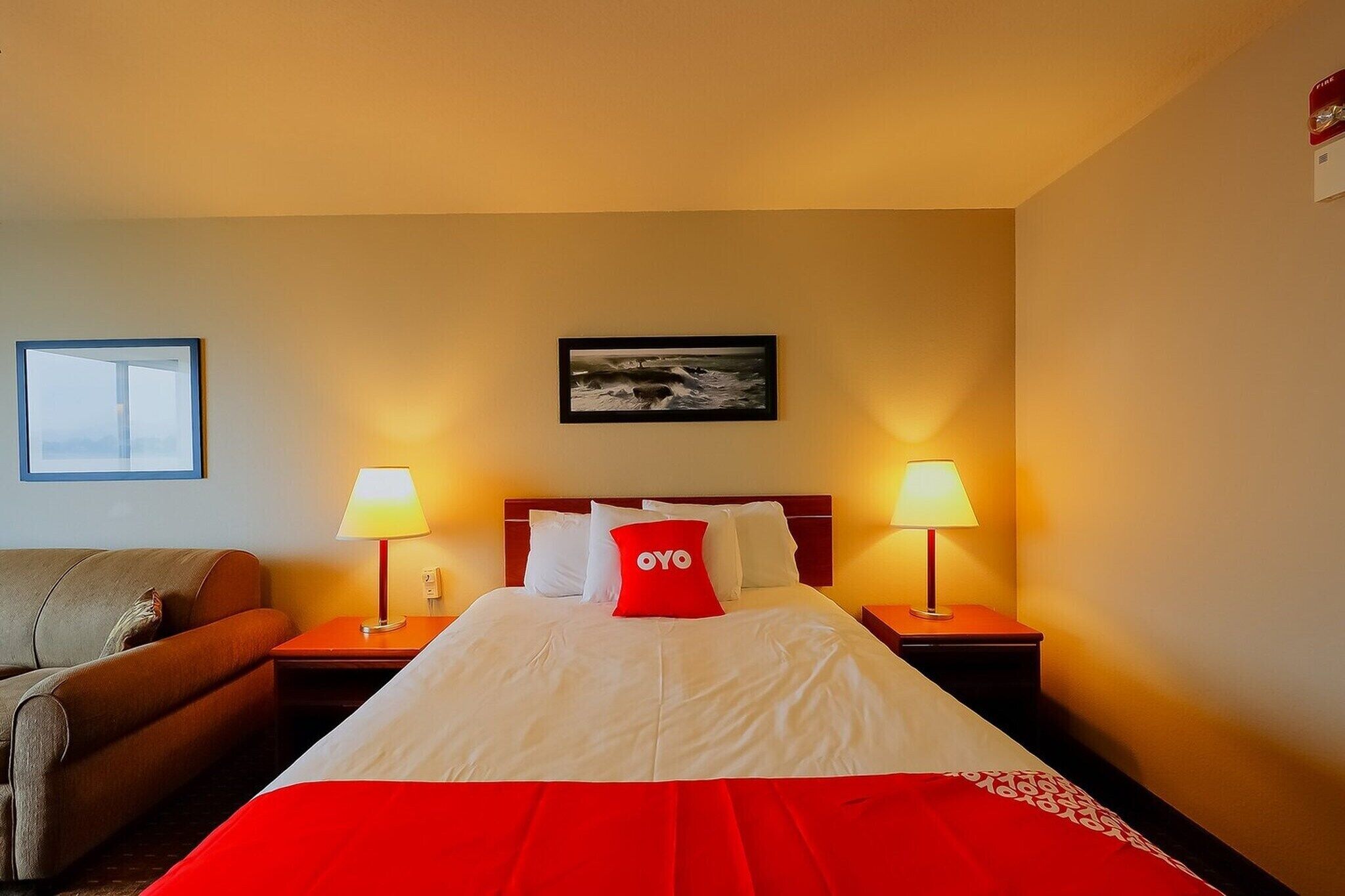 Une chambre d’hôtel avec un lit, deux lampes de chevet, un oreiller rouge, un tableau encadré au mur et un canapé.