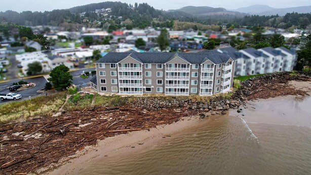 A multi-story hotel with a flat roof, located near a beach with driftwood and a stone wall.