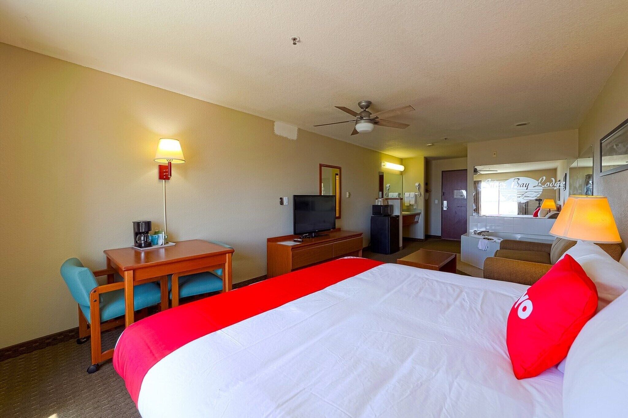 Ein Hotelzimmer mit einem Bett, einem Schreibtisch mit Stuhl, einem Fernseher, einem Deckenventilator und einem durchgegangene Tür sichtbaren Badezimmer.