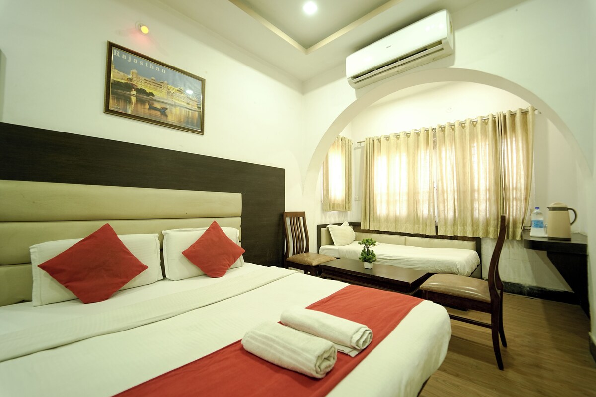 A hotel room with a large bed, a smaller bed, a desk, a chair, an air conditioning unit, and a framed picture on the wall.