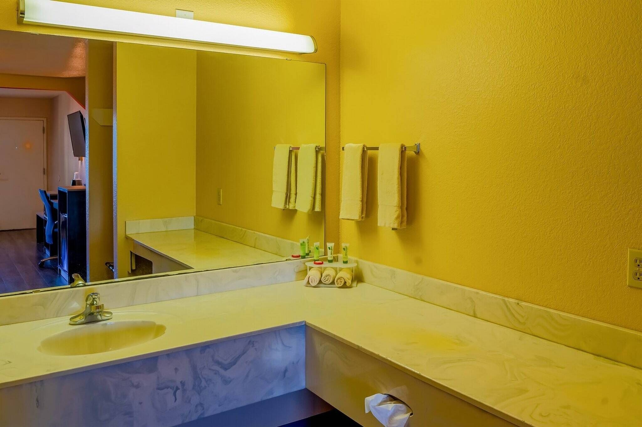 A bathroom with a yellow wall, a large mirror, a sink, and towels hanging on a rack.