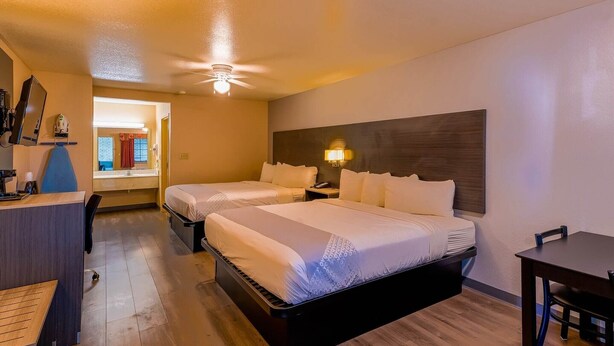 A hotel room with two beds, a desk, a chair, a ceiling fan, and a nightstand with a lamp.