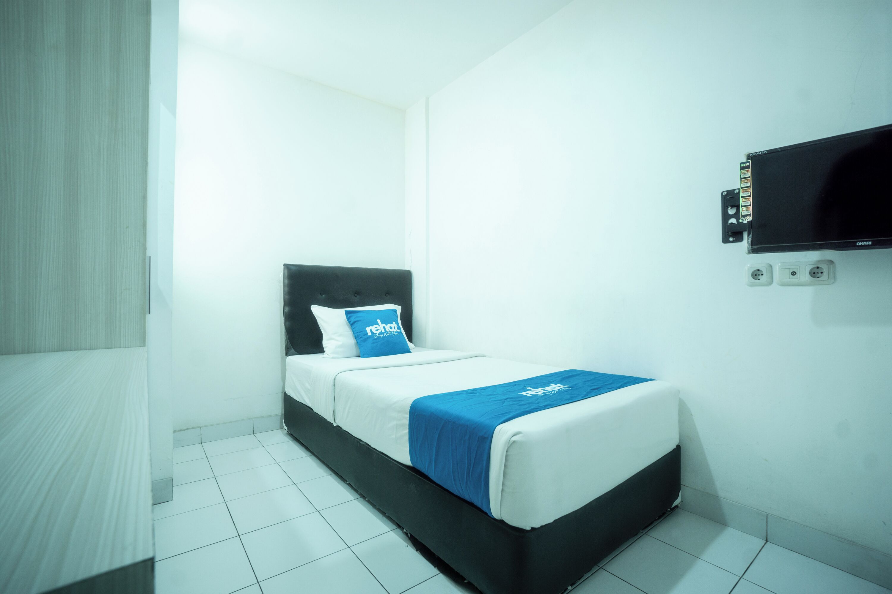 A single bed with a black headboard, white bedding, and a blue accent pillow. There is a flat-screen TV mounted on the wall to the right of the bed.