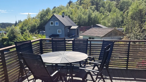 Une terrasse en bois avec une table ronde et des chaises, entourée d’une clôture en bois, donnant sur un quartier résidentiel avec des maisons et des arbres.