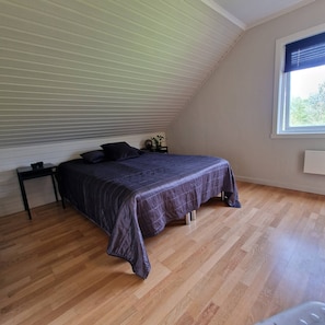Ett sovrum med ett sluttande tak, en säng med ett mörkt täcke, ett litet sängbord och ett fönster med persienner.