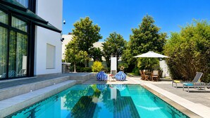 A swimming pool with lounge chairs and umbrellas, surrounded by trees and a modern building. - Villa Solis - Elegant Villa with Private Pool (Marsa)