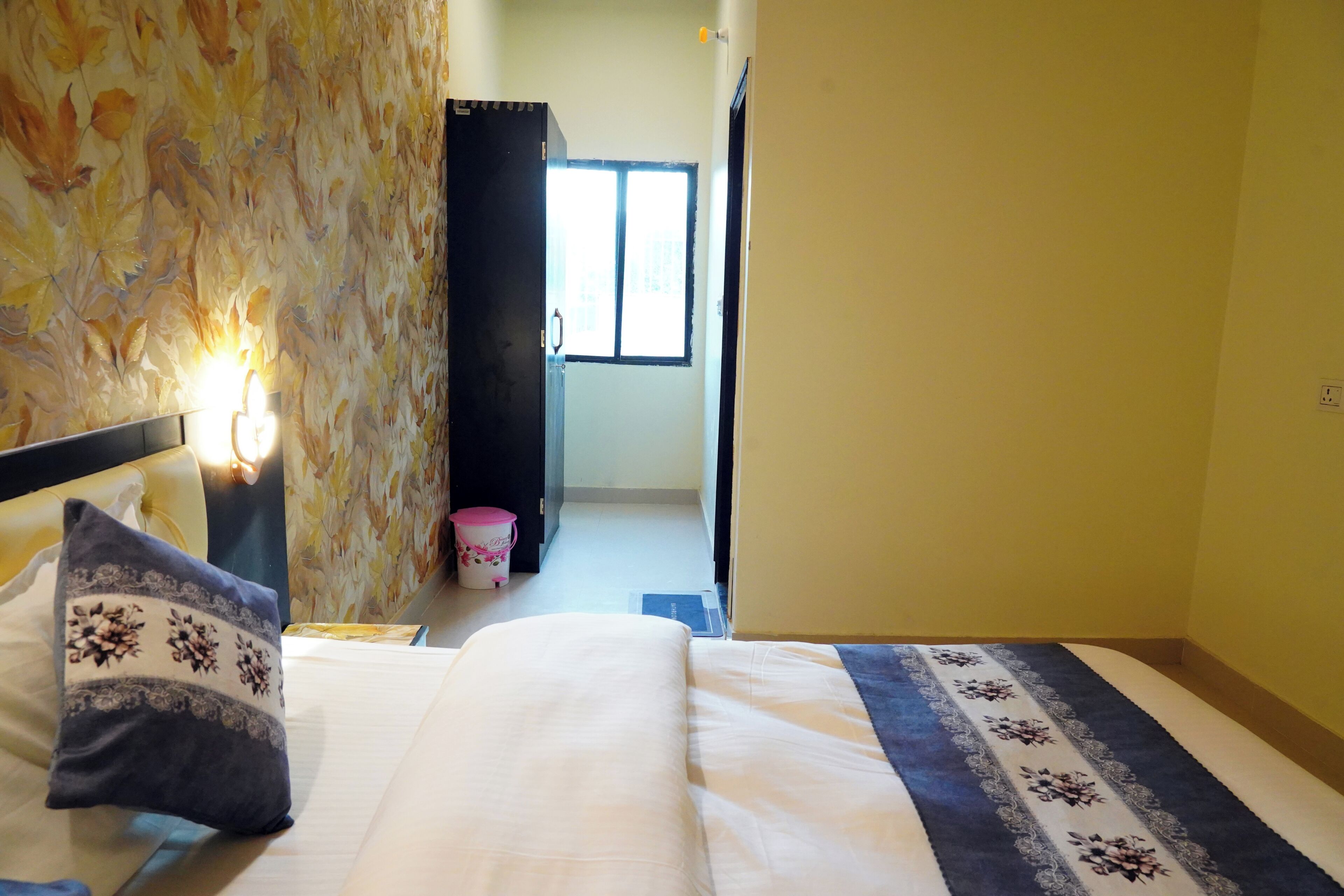 Habitación de hotel con cama, mesita de noche con lámpara, ventana y puerta que da a otra habitación.