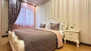Quarto com uma cama grande, cabeceira ornamentada, mesa de cabeceira e janela com cortinas.