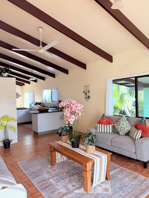 A living room with a sofa, a wooden coffee table, a hanging plant, and a kitchen in the background. - Mountain Views, 3BR Family Home, Near Muri Beach (Takitumu District)