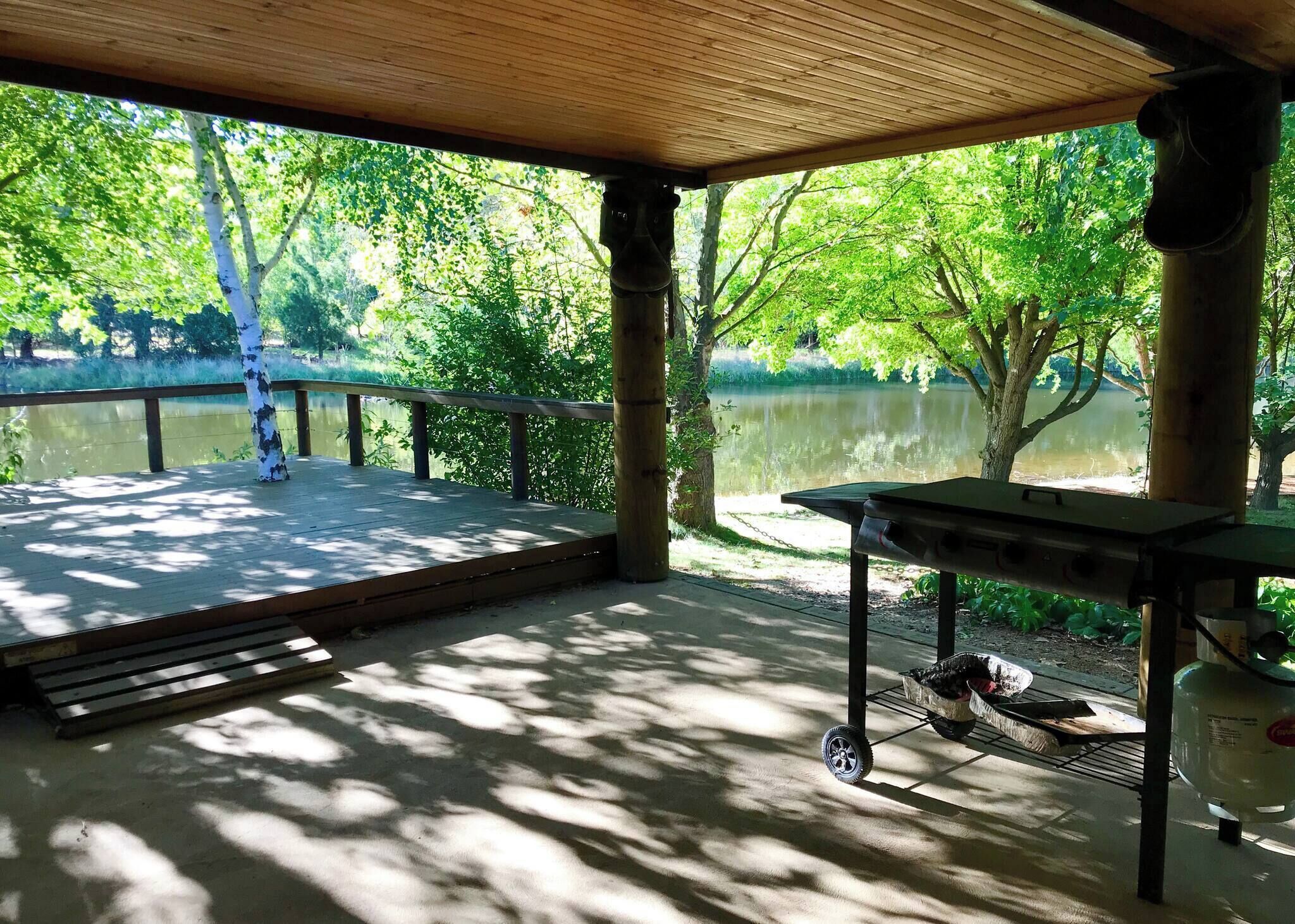 Un espacio al aire libre techado con una terraza de madera, una mesa con una silla y vistas a un lago rodeado de árboles.