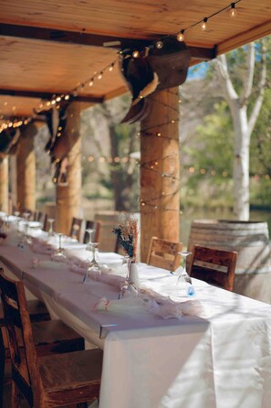 An outdoor dining area with a long table set for a meal, string lights, and rustic wooden pillars.