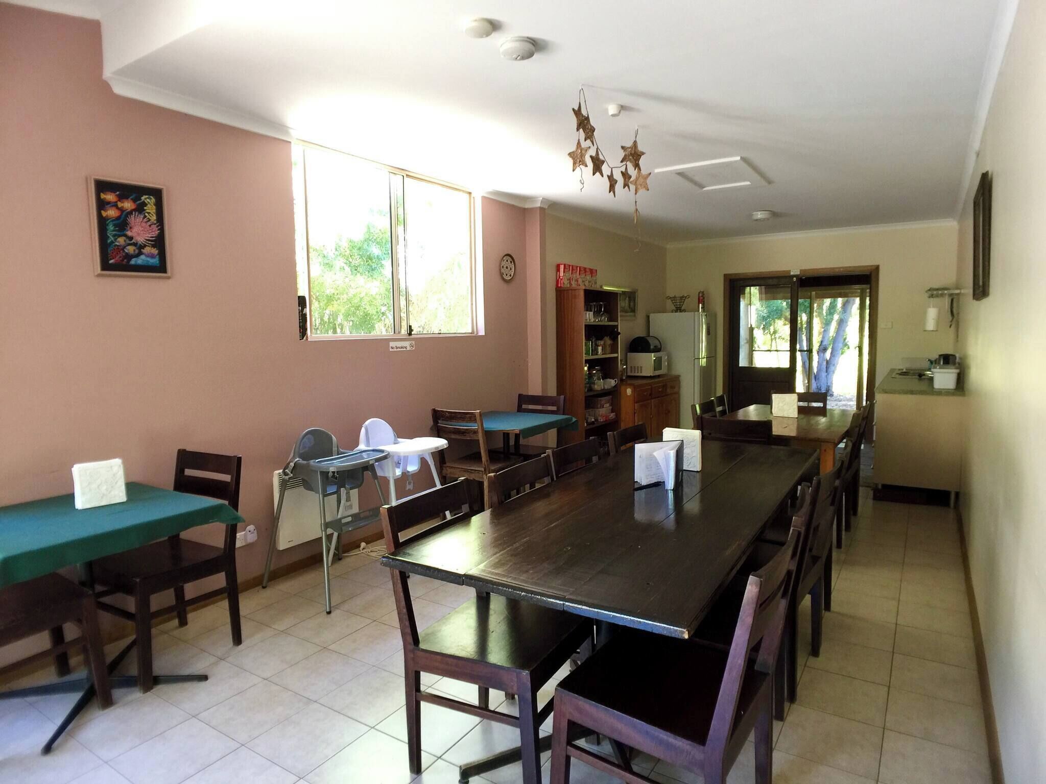 A dining area with a long wooden table, chairs, a small kitchen area, and a window with a view of trees.