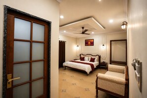 A bedroom with a bed, a chair, a ceiling fan, a nightstand, and a mirror. - Vaanam One (Ujjain)