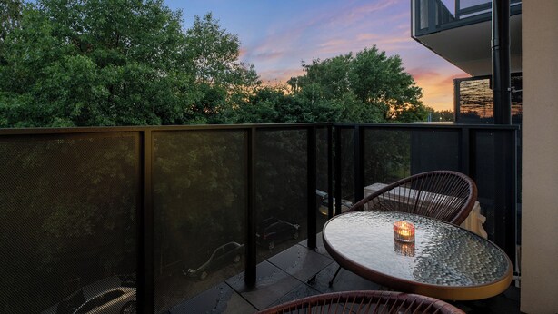 Ein Balkon mit Glastisch und Stühlen, Blick auf Bäume und Sonnenuntergang.