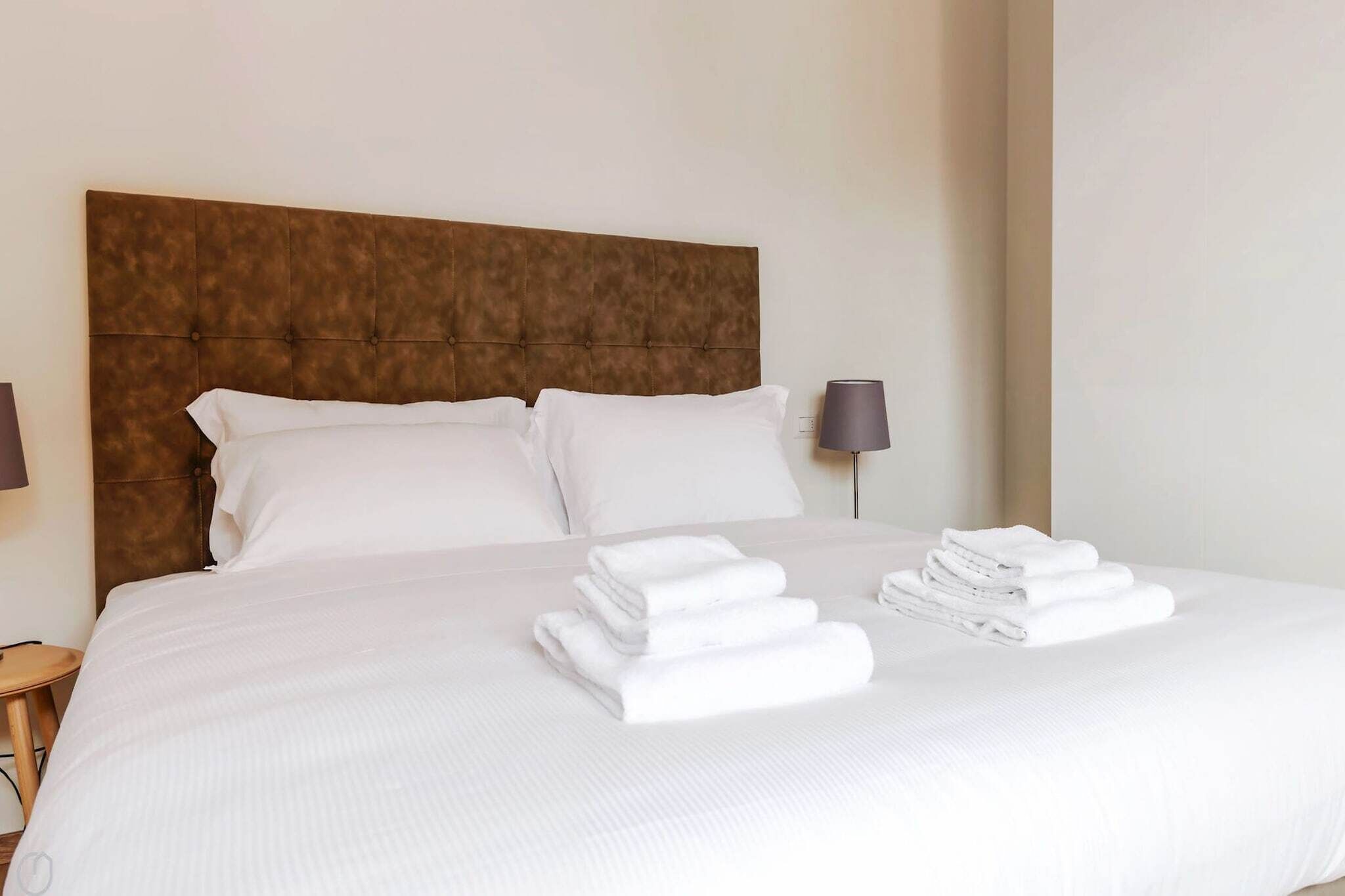 Una cama bien hecha con un cabecero marrón, almohadas blancas y una pila de toallas blancas dobladas.