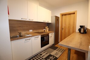 Une cuisine compacte avec des meubles blancs, un plan de travail en bois, un four encastré, un réfrigérateur et une machine à café.