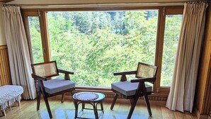 Ein Zimmer mit einem Bett, zwei Stühlen, einem kleinen Tisch und großen Fenstern mit Blick auf Grünflächen.
