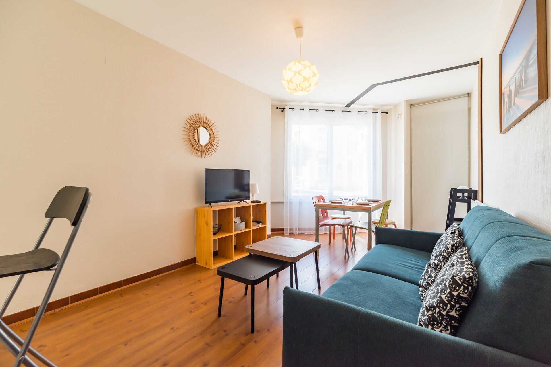 A living room with a sofa, a TV, a wooden TV stand, a dining table with chairs, and a window with sheer curtains.