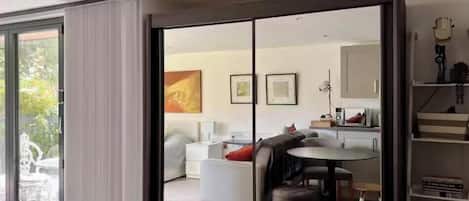 Una sala de estar moderna con un gran ventanal corredizo que da a un balcón, un comedor con una mesa redonda y una cocina visible a través del vidrio.