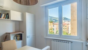 A modern room with a white table, a chair, a bookshelf, a white cabinet, and a window with a view of buildings. - Casa Vacanze Ca Zigoela (La Spezia)