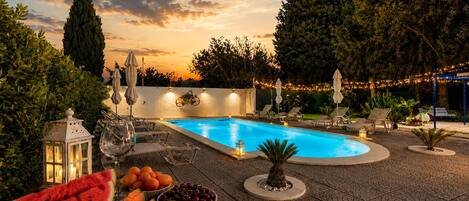 An outdoor swimming pool area with lounge chairs, a fruit bowl, and decorative lighting.