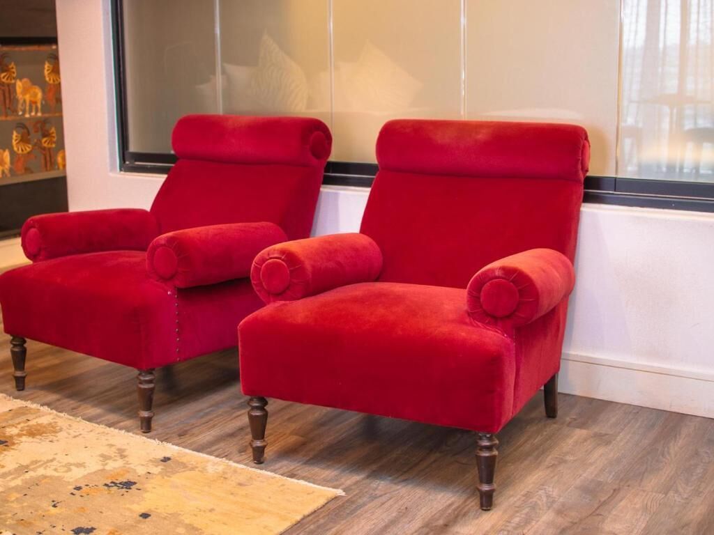 Two red velvet armchairs with wooden legs, positioned in front of a window with a view of a building.
