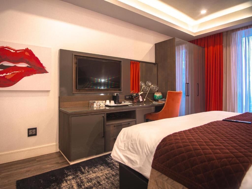 Quarto de hotel com cama, escrivaninha, computador, televisão e uma janela grande com cortinas.