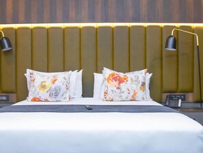 A hotel room with a bed, two pillows with floral patterns, a bedside table with a phone, and a wall-mounted lamp.