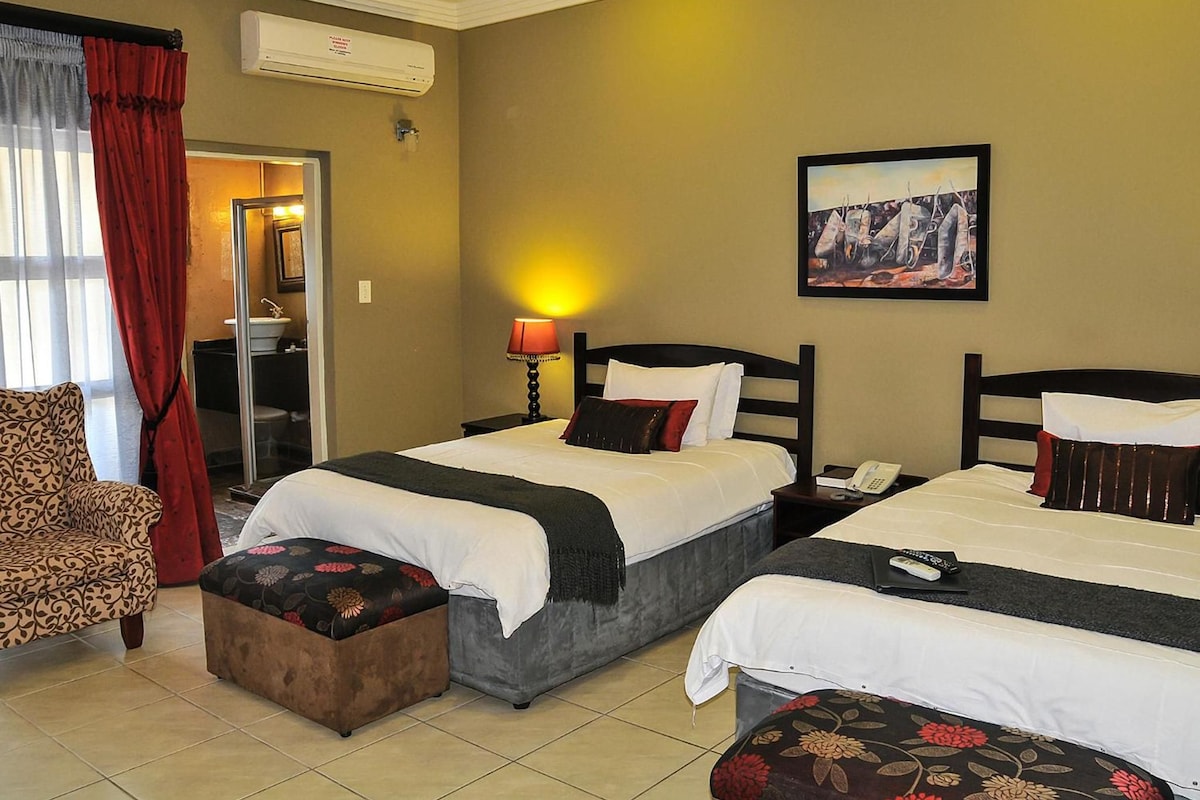 Habitación de hotel con dos camas, un sofá, una mesita, una lámpara y un cuadro en la pared.