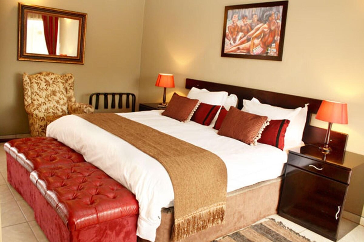 A hotel room with a large bed, a red upholstered bench, two bedside tables with lamps, a chair, and a painting on the wall.