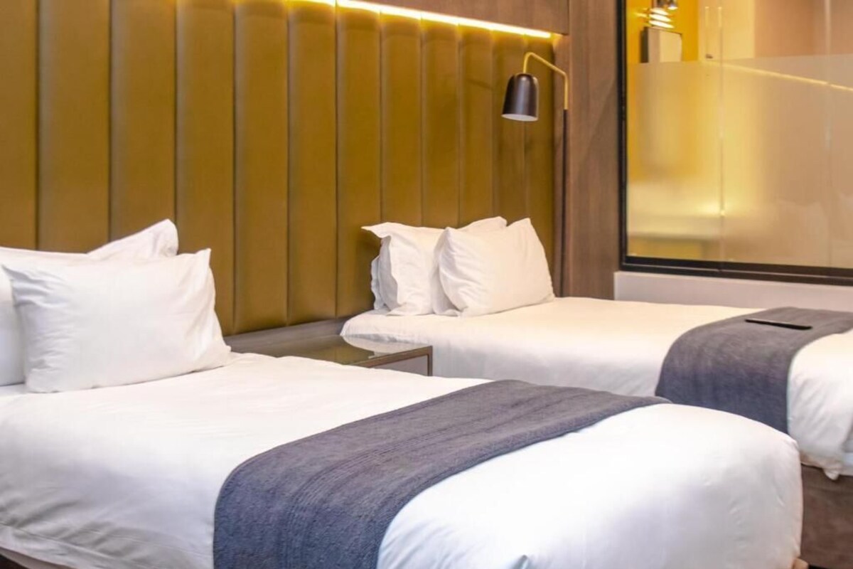 Two single beds with white linens and a gray blanket, a bedside table, and a wall-mounted lamp.